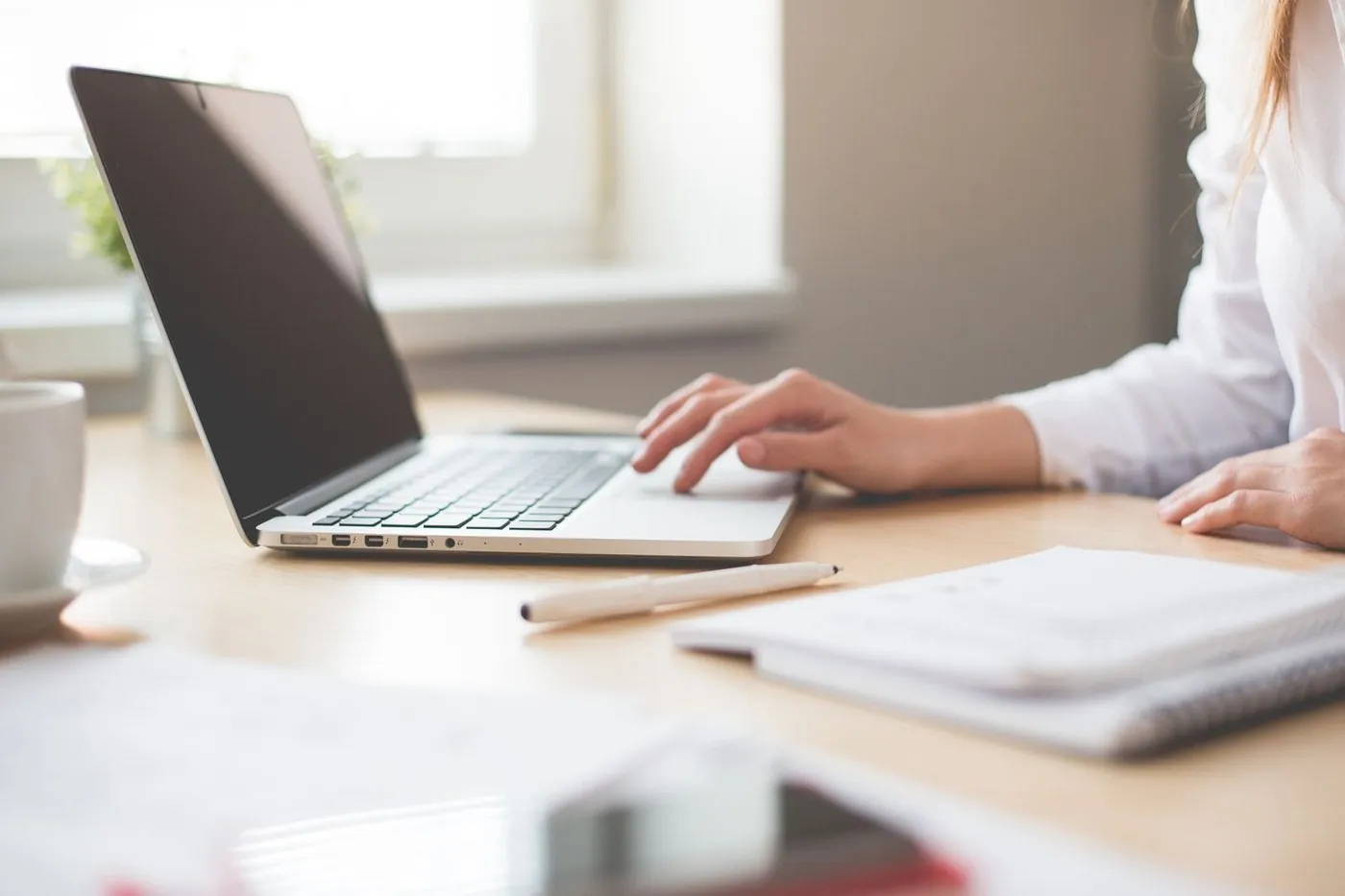 Hands on a silver laptop beside a spiral notebook on a wooden desk.