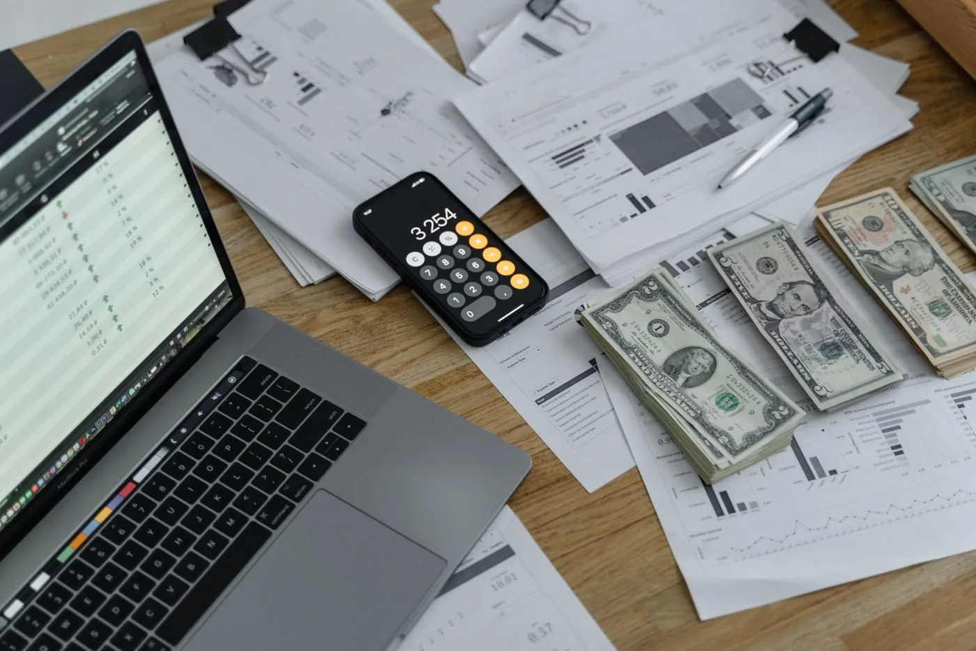 A wooden desk with a laptop, stacks of United States bills, a phone calculator, and papers.