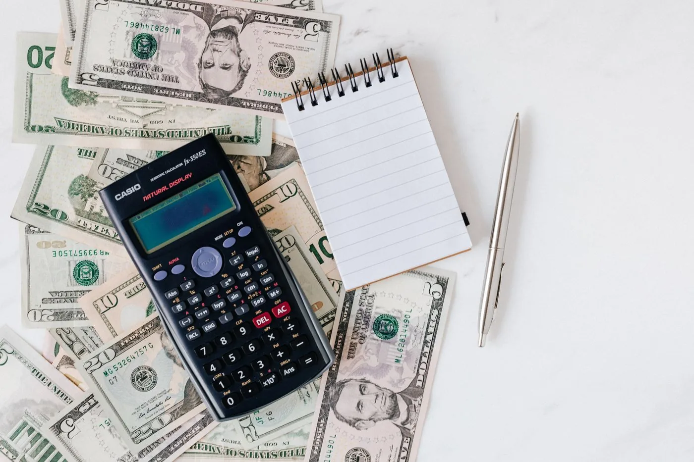 A calculator, notepad, and pen on a pile of United States bills